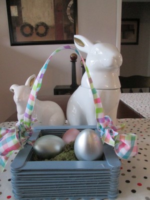 Craft Stick Basket - blue basket with spring colored checkered ribbon handle and pearlized eggs inside, sitting in front of two white ceramic rabbits