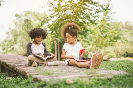 Two kids sitting on a rustic wooden bridge, reading books.