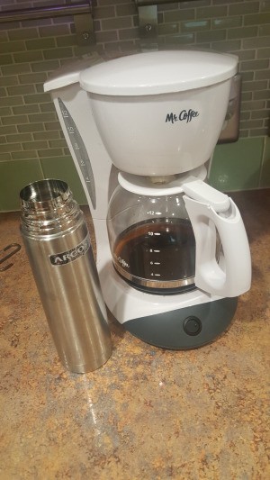 A coffee pot next to a stainless steel travel mug.