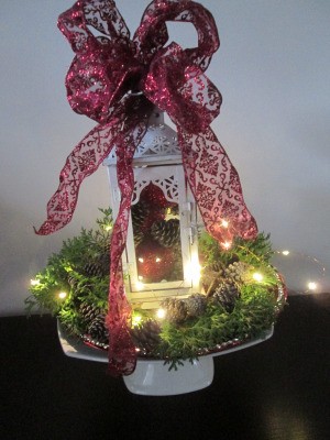 Making A Lantern Part Of Your Holiday Decor - ornament filled white lantern on a display stand surrounded by greenery, pine cones, and fairy lights