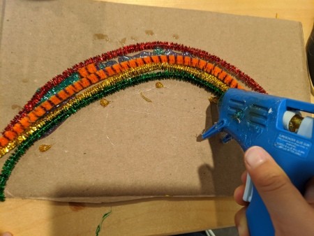 Rainbow Name Room Decor - child glueing pipe cleaners to cardboard with hot glue gun