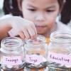 A child placing coins into three jars, marked savings, toys and education.
