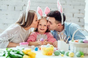 A man, woman and child with bunny ears on, decorating eggs.