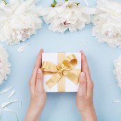 Hands holding a gift in a white box with a gold bow, surrounded by white flowers.