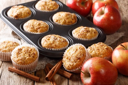 A muffin tin with apple cinnamon muffins inside, next to apples and cinnamon sticks.