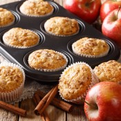 A muffin tin with apple cinnamon muffins inside, next to apples and cinnamon sticks.