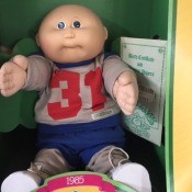 A Cabbage Patch doll in the box wearing a grey sweatshirt with the number 31