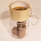 A roll of tape on top of a recycled glass jar containing a roll of twine.