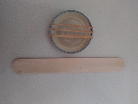 A bottlecap with three rubber bands and a craft stick.