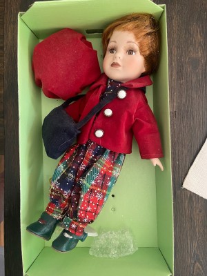 A porcelain doll in the box, with a red hat and coat.