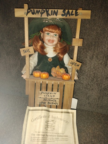 A porcelain doll selling pumpkins at a stand.