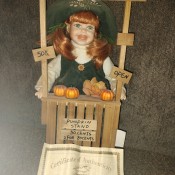 A porcelain doll selling pumpkins at a stand.